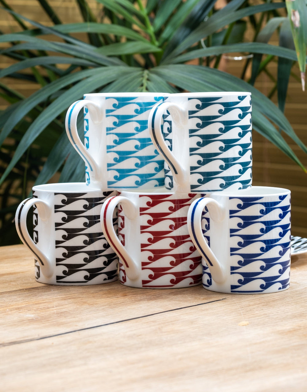 Five wave patterned mugs stacked on a wooden surface with green plants in the background