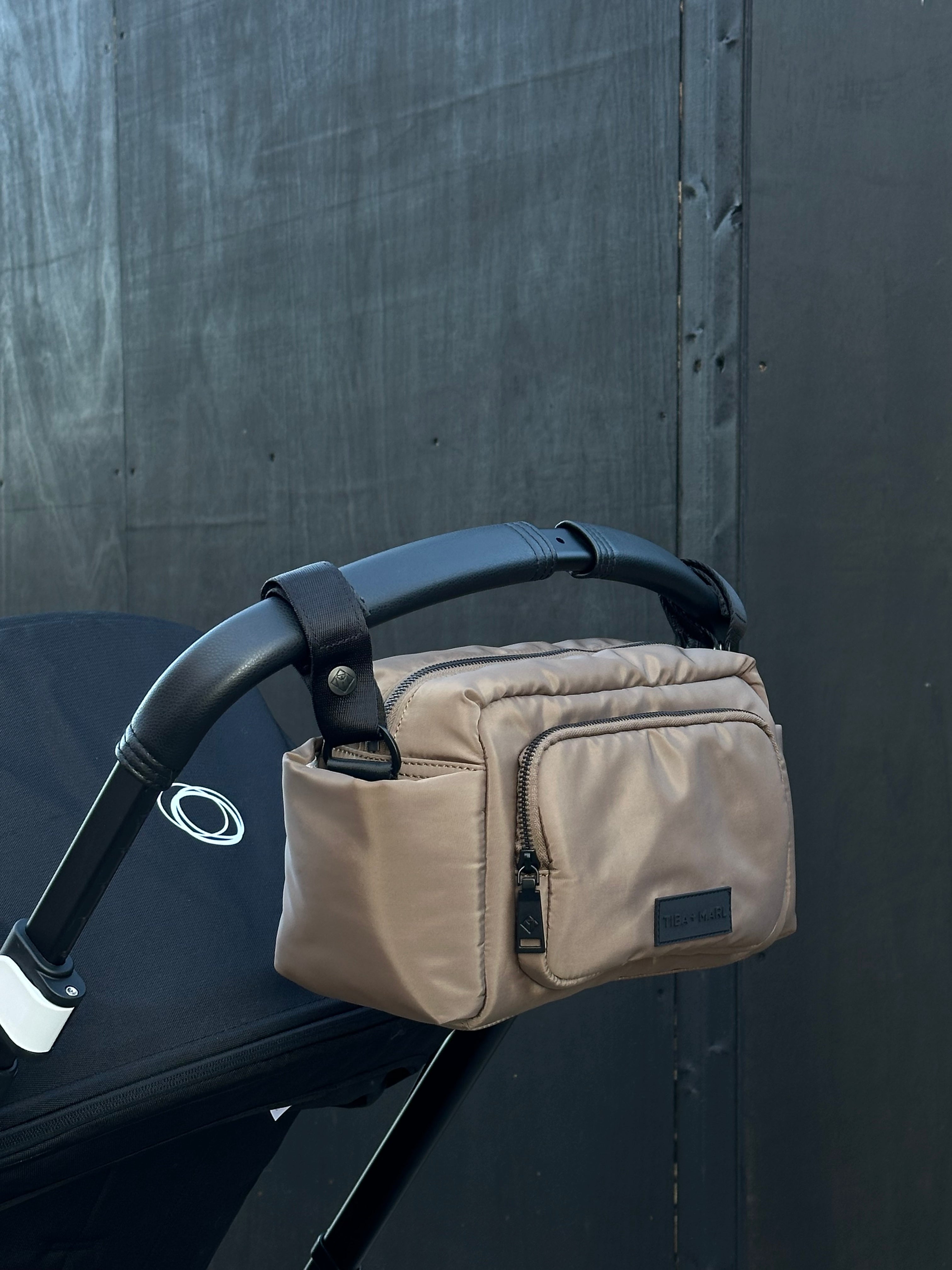 Beige diaper bag attached to a black stroller against a dark background
