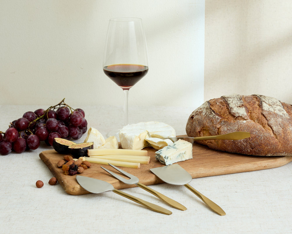 Wooden board with cheese, bread, grapes, and a glass of red wine on a white surface.