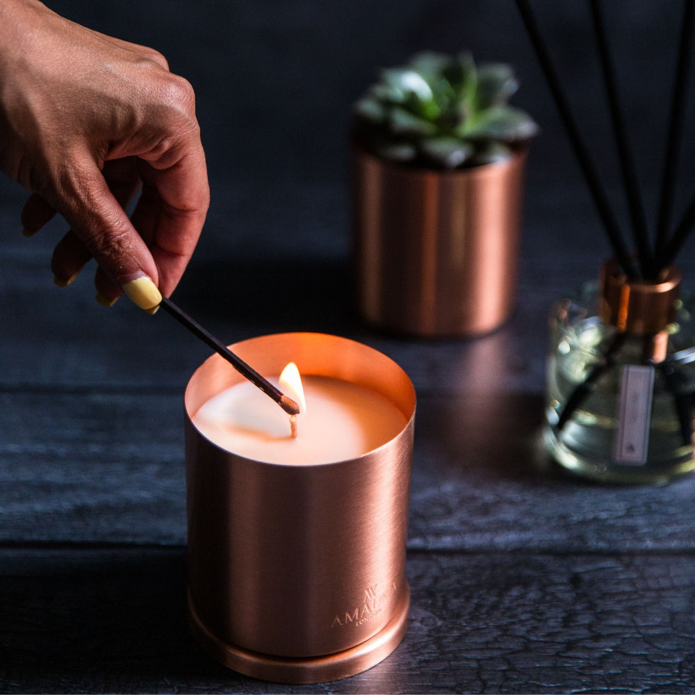 Hand lighting a candle in a copper holder with a plant and diffuser in the background.
