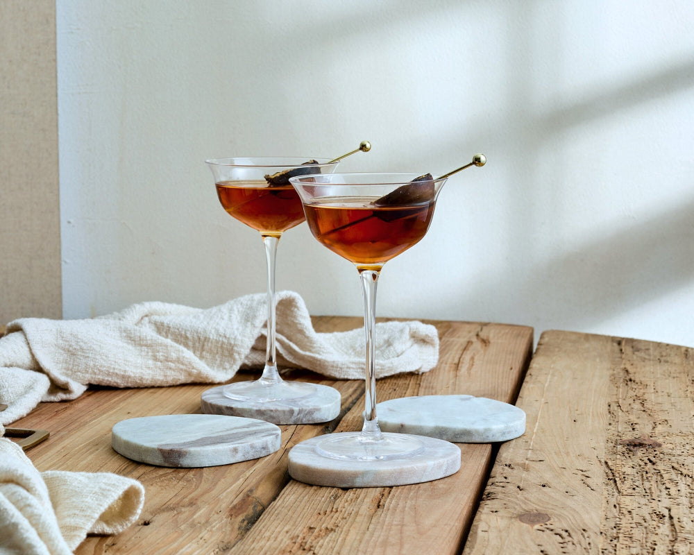 Two cocktails in martini glasses on a wooden table with marble coasters.