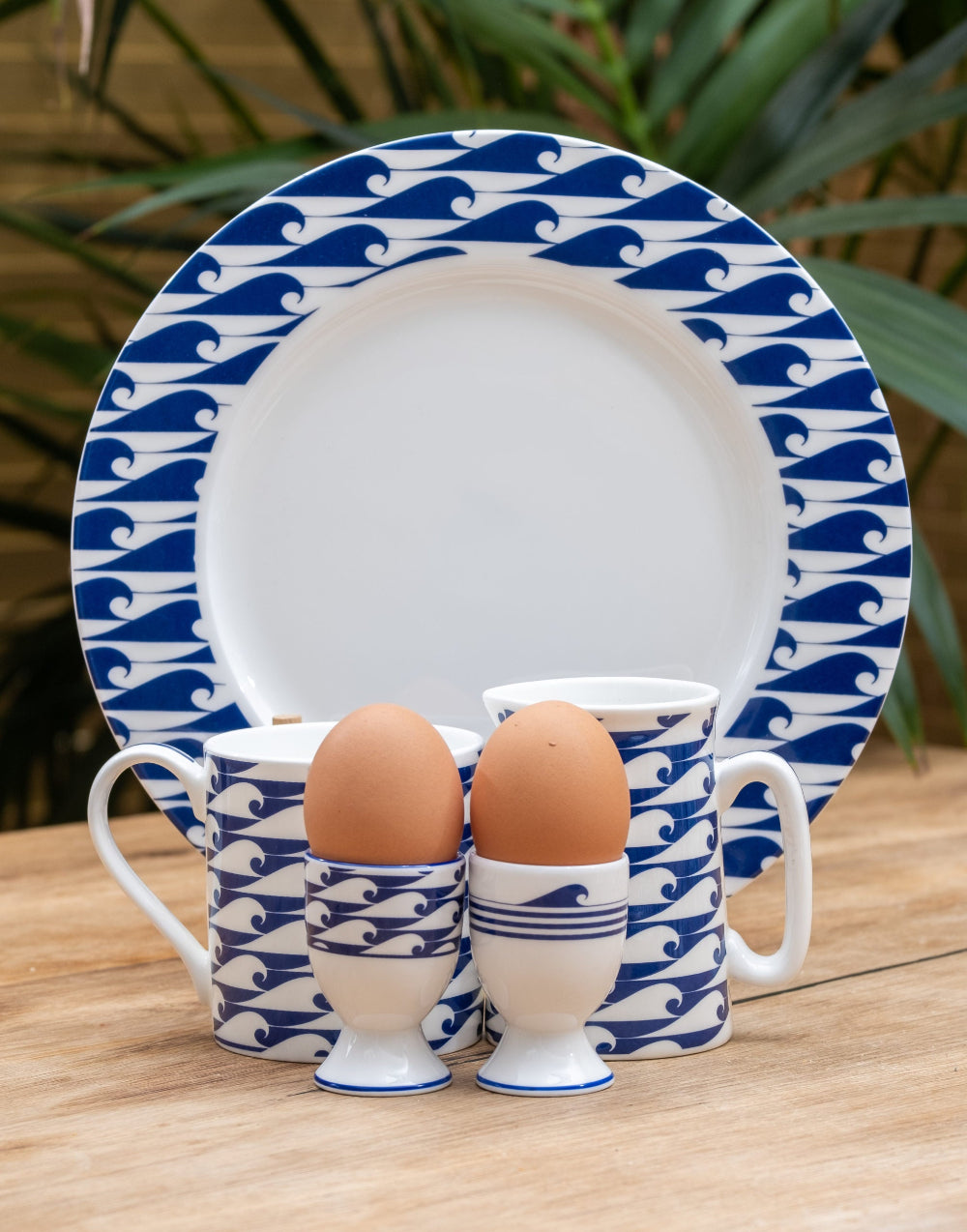 Ceramic set with blue wave pattern including a plate, mug, and egg cups on a wooden surface.