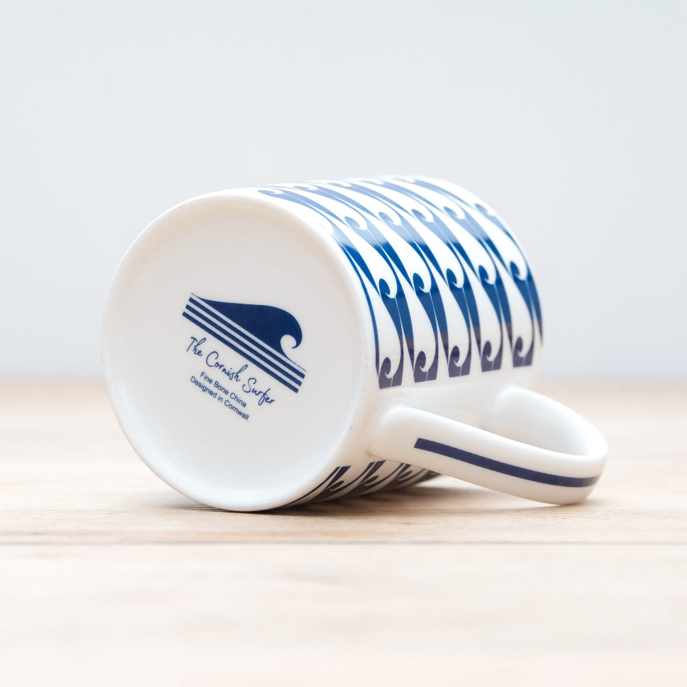 The Cornish Surfer logo in blue on the bottom of a mug on its side laying on a wooden table