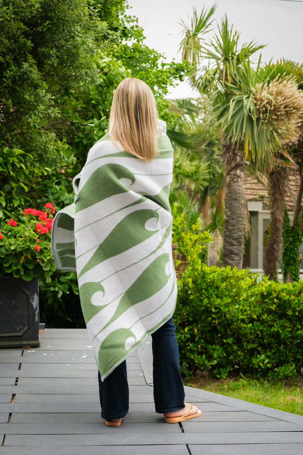 Person wrapped in a green and white patterned blanket standing on a wooden deck with greenery in the background