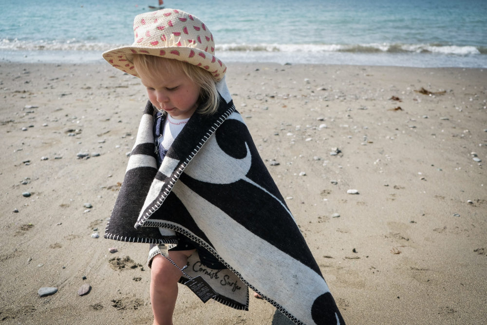 Child on a beach wearing a black and white patterned blanket and sun hat.