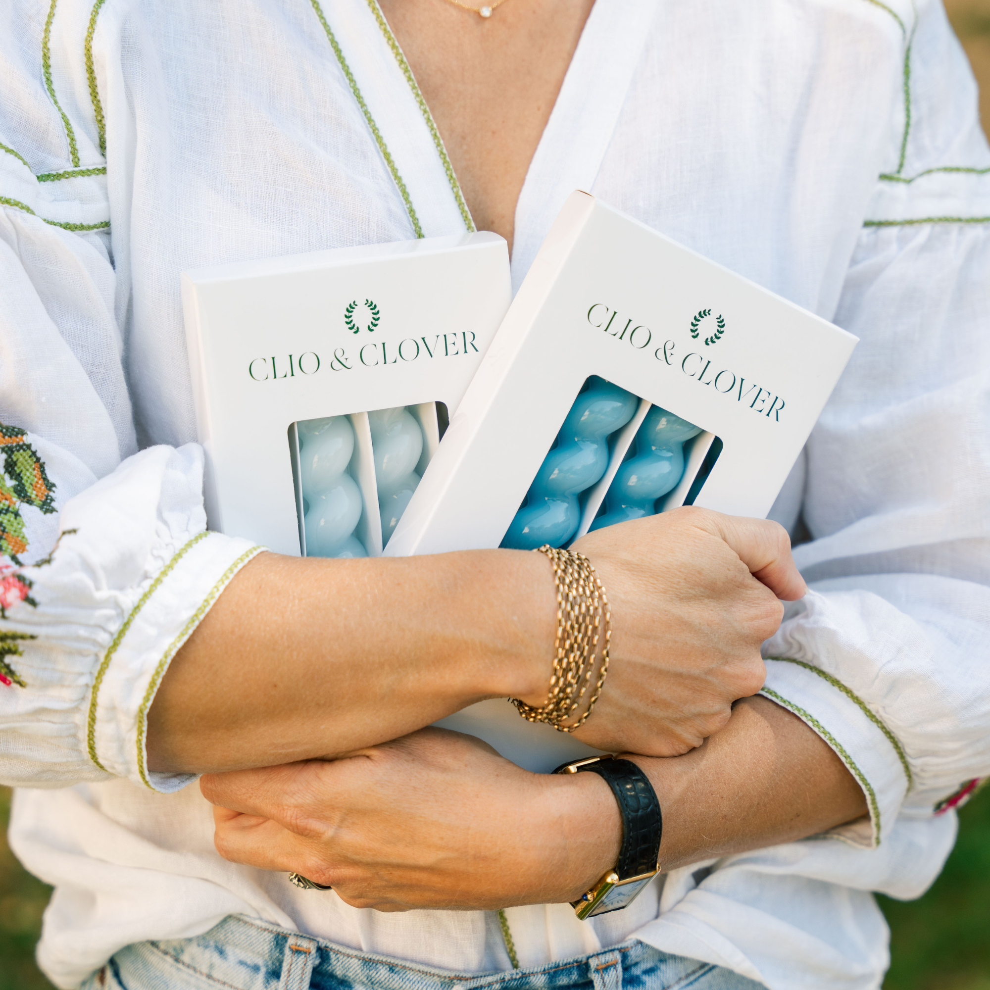 Person holding two boxes labeled 'Clio & Clover' with a white embroidered garment. Baby Blue and Blue Taper candles spiral design.