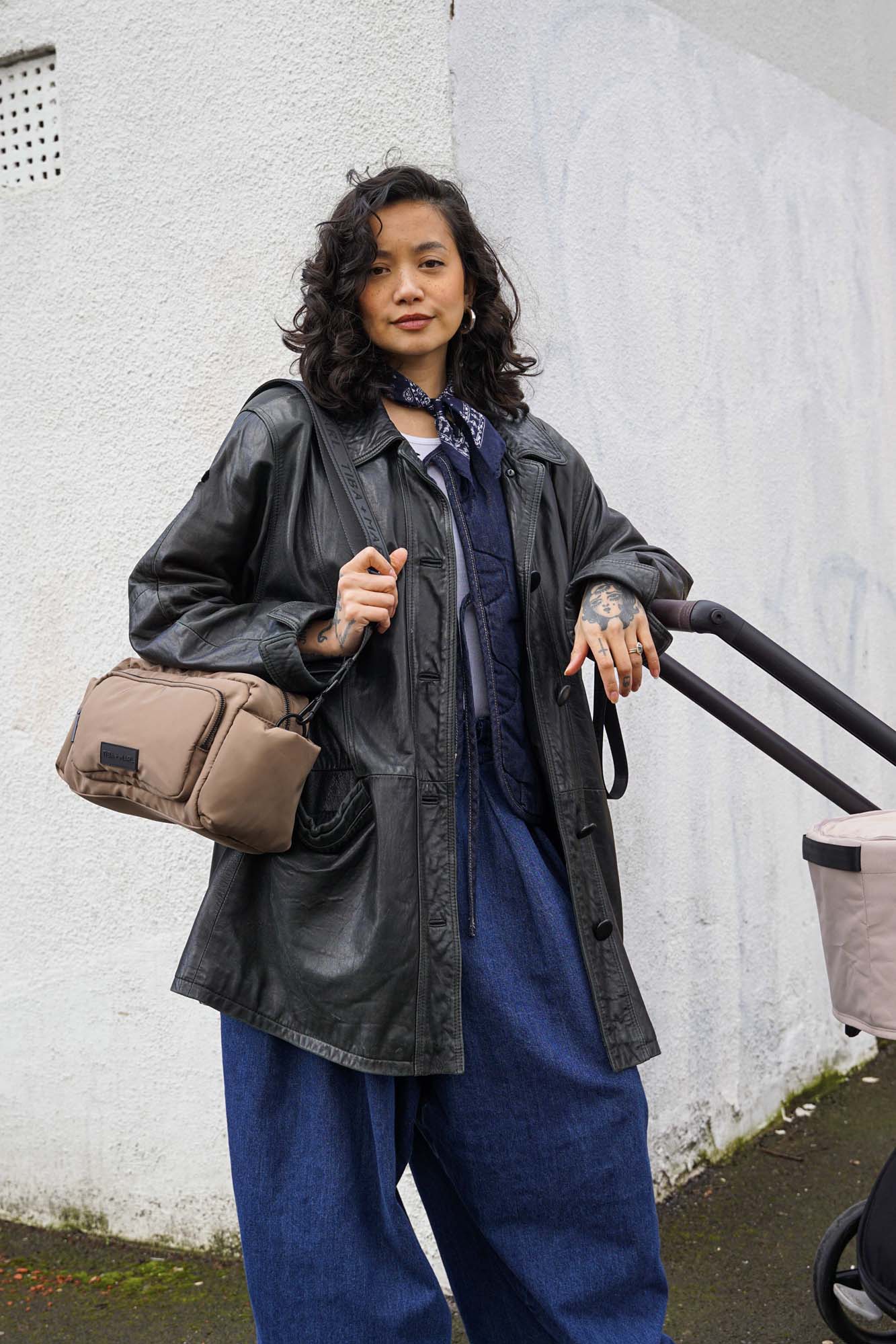 Person wearing a black coat and blue pants holding a beige bag against a white wall. Leaning on a baby buggy handle.