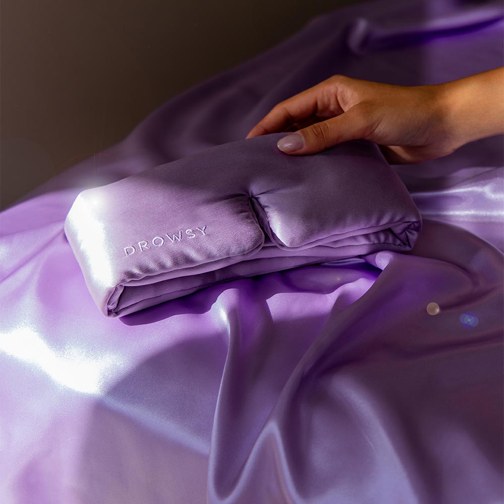 Folded purple sleep mask being held by a hand on a purple fabric background.