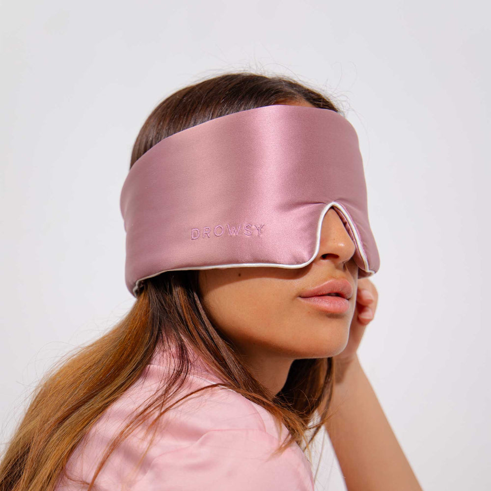 A person wearing a pink silk sleep mask with white piping detail along the edges.