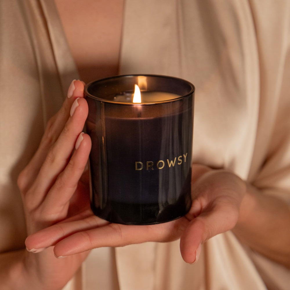 Person holding a lit Drowsy candle in a dark glass container.