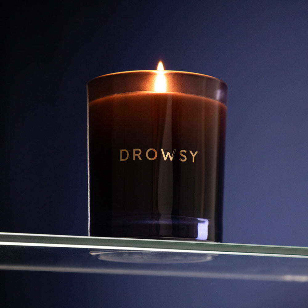 Drowsy candle in a dark glass container on a reflective surface with a dark purple background