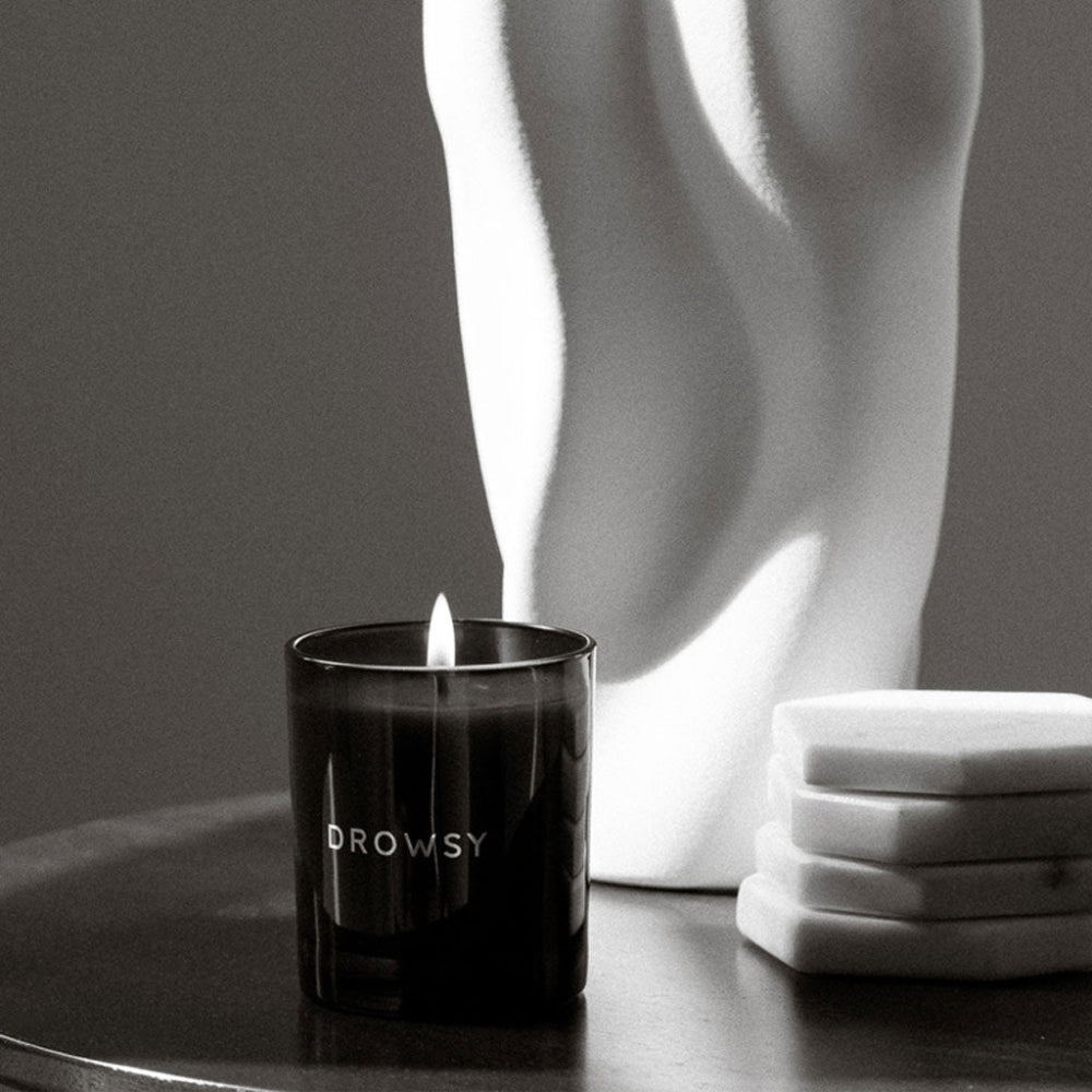 Black candle labelled 'Drowsy' on a table with a white abstract sculpture in the background.