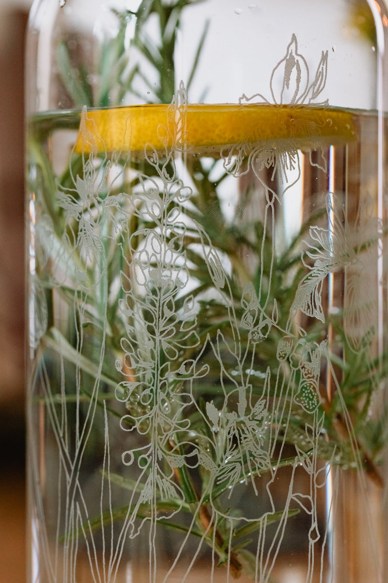 Glass jar with engraved plant design and slice on lemon in water containing herbs.