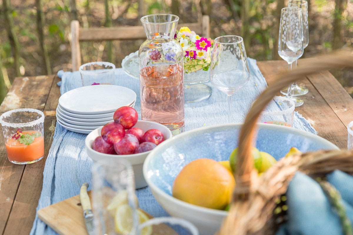 Outdoor table setting with fruits, glasses, and a basket on a wooden table.