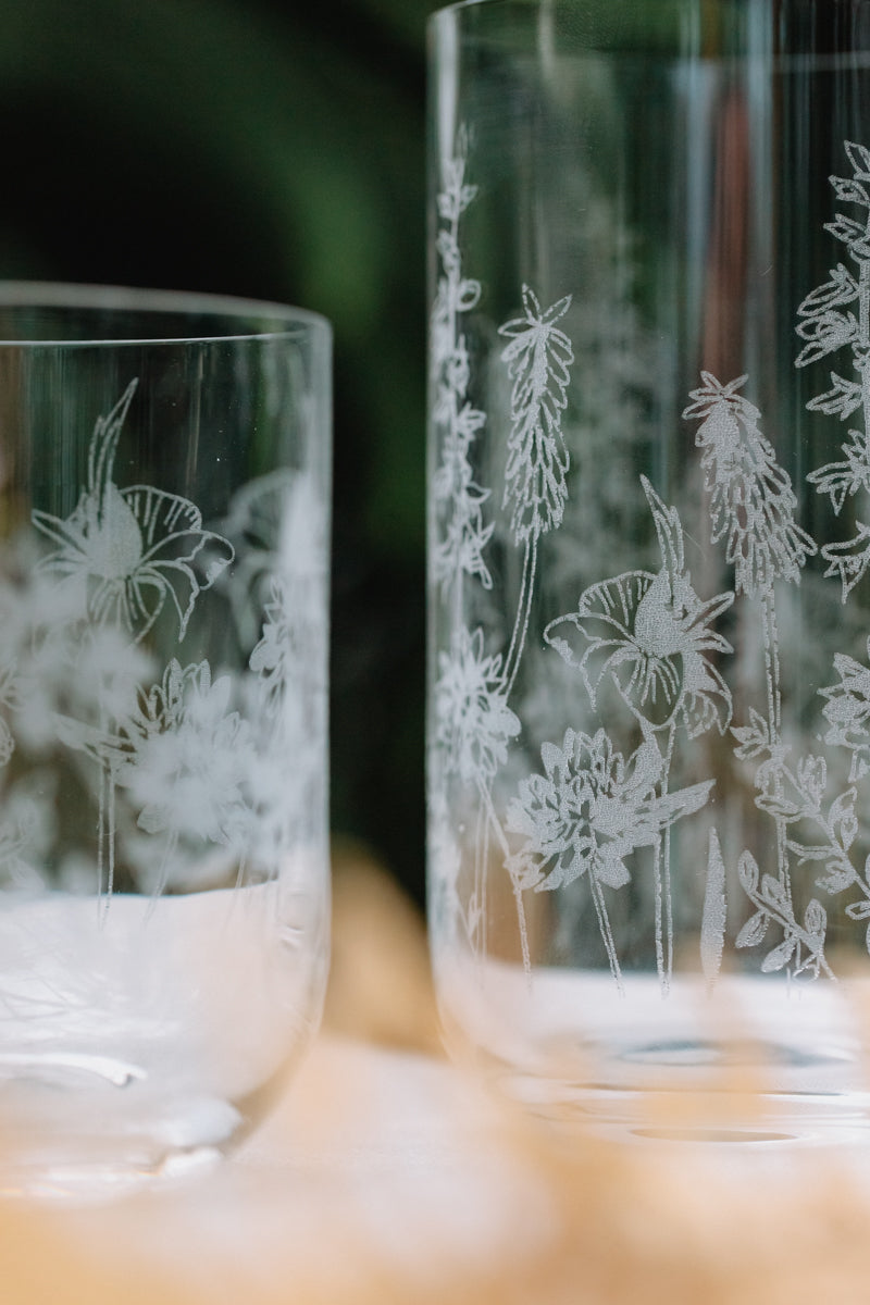 Two etched glass tumblers with floral and butterfly designs on a blurred background.