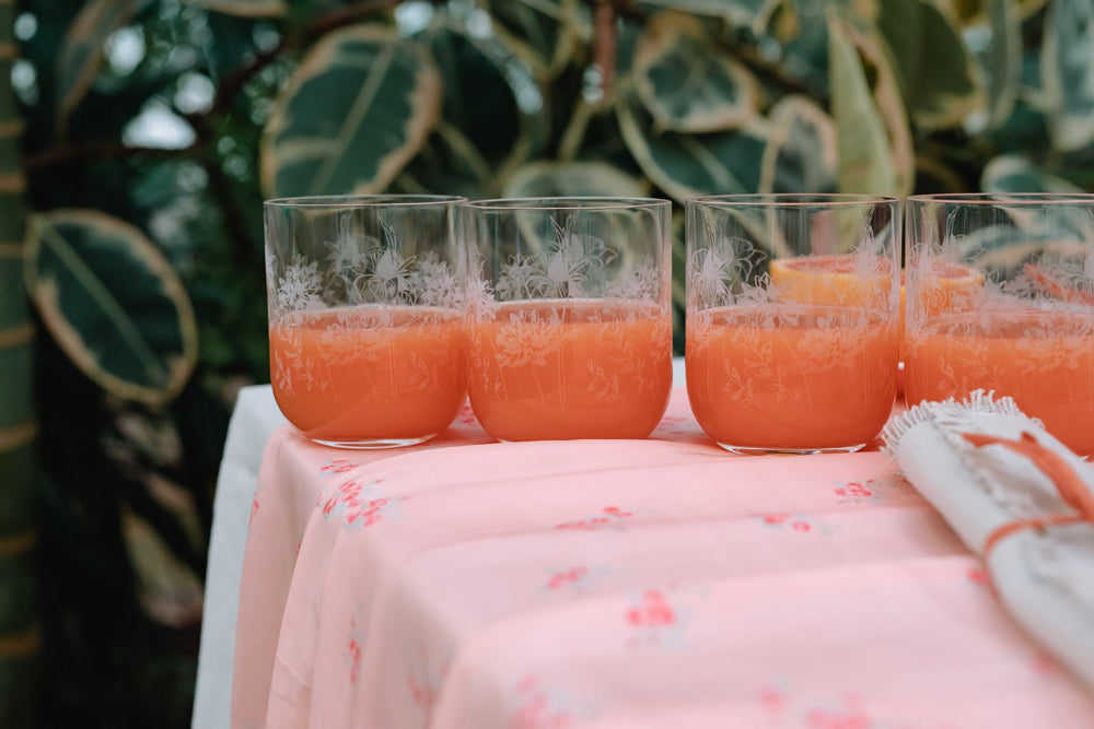 Four glasses of orange juice on a table with a floral tablecloth, surrounded by green foliage.