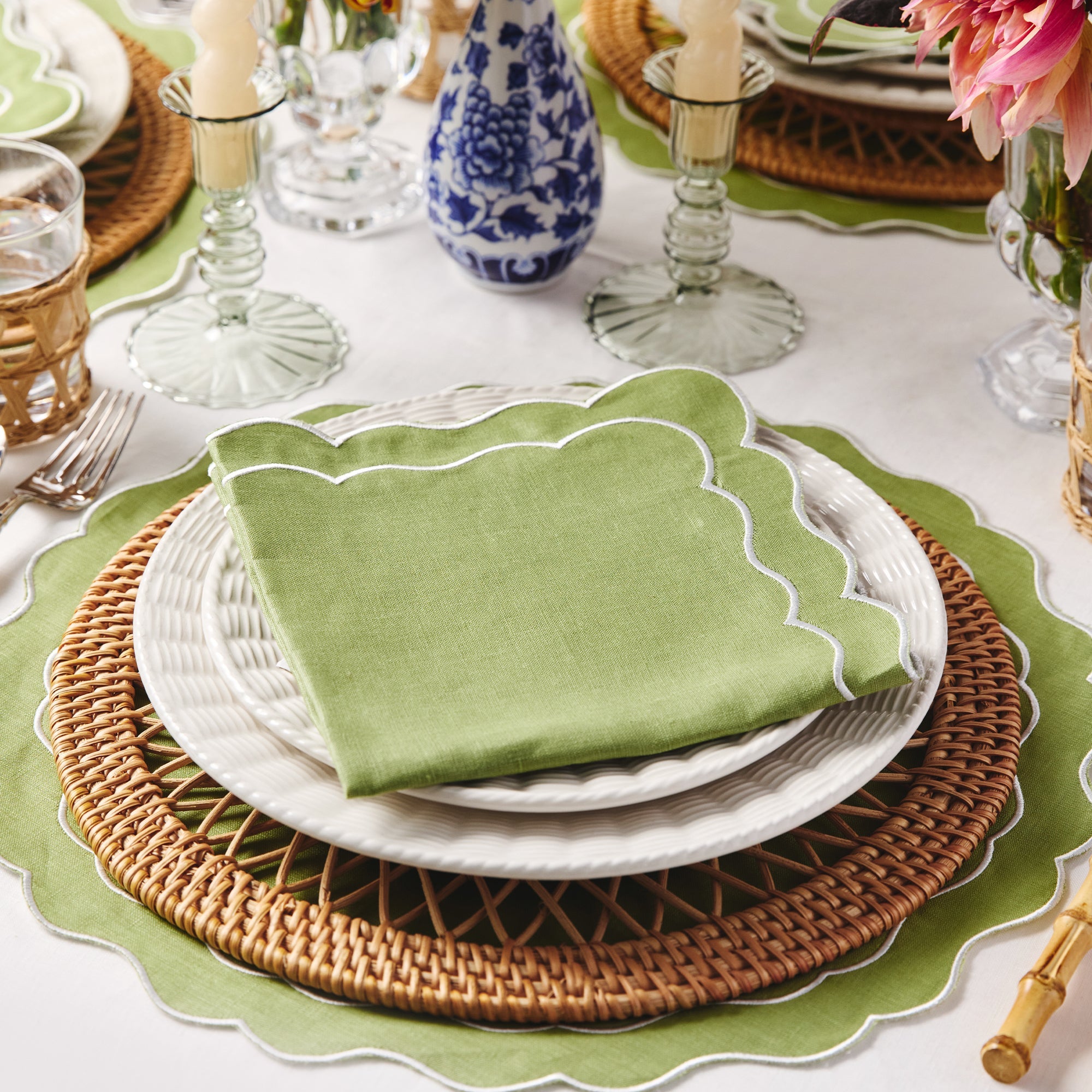 Elegant table setting with green napkin, white plates, and rattan placemats on a white tablecloth. Blue and white decorative vase.