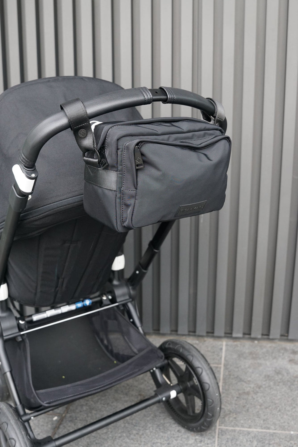 Black stroller with a bag attached against a corregated metal wall.