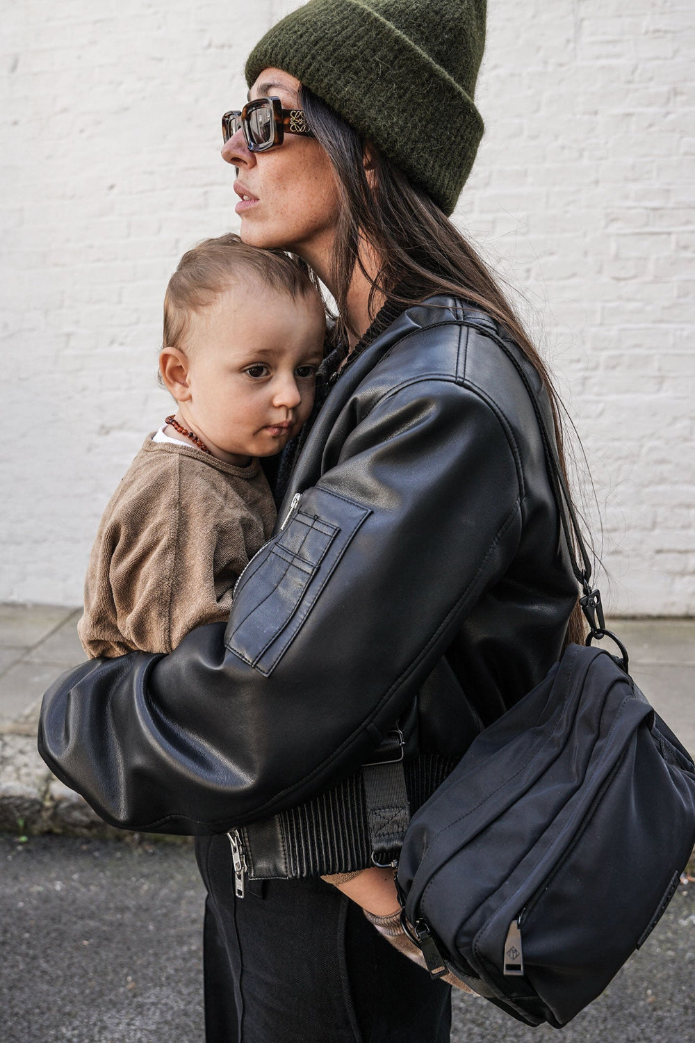 Woman in black leather jacket holding a child, wearing sunglasses, black crossbody bag and a green beanie, against a white brick wall.