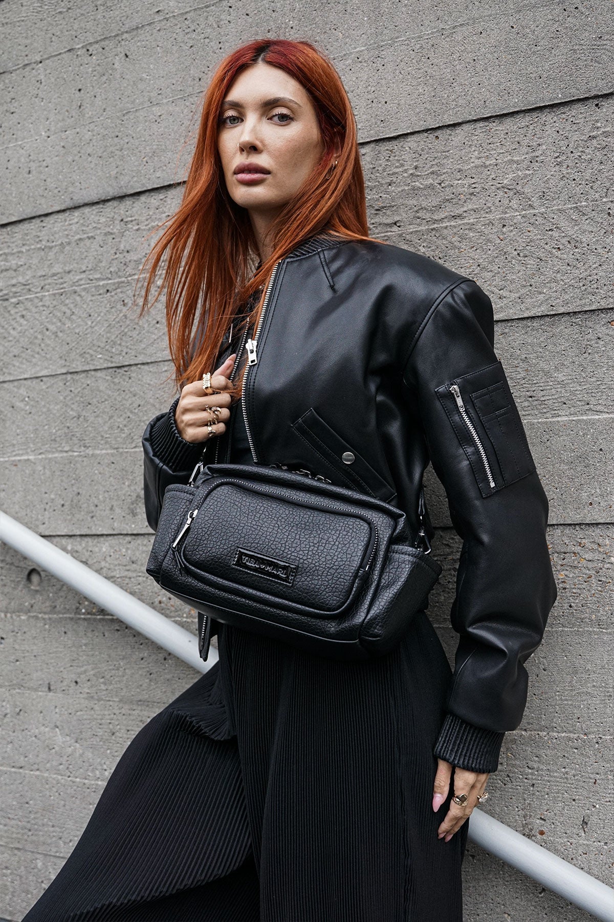 Red haired Woman wearing a black leather jacket and carrying a black crossbody bag against a grey wall.