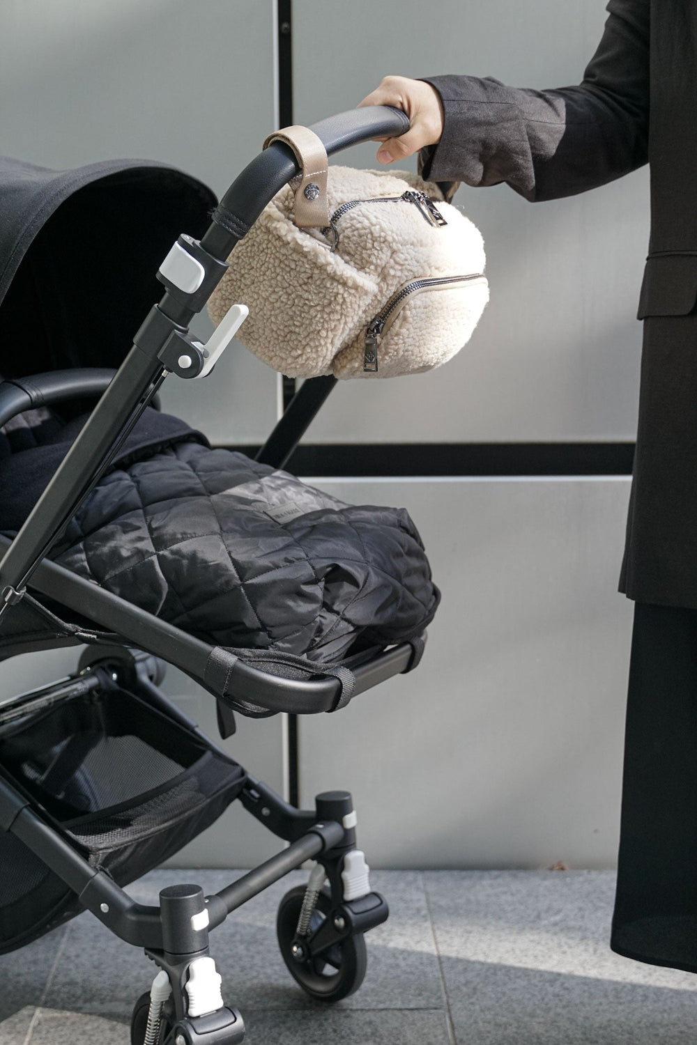 Person holding a stroller with a fleece beige bag handle clipped to buggy handle against a neutral background