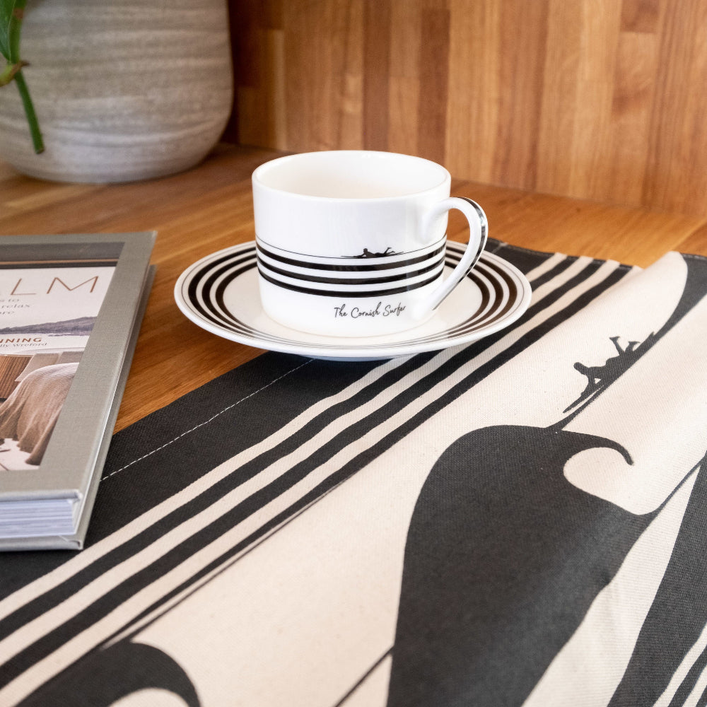 White cup with black stripes on a saucer on a table with a matching tea towel