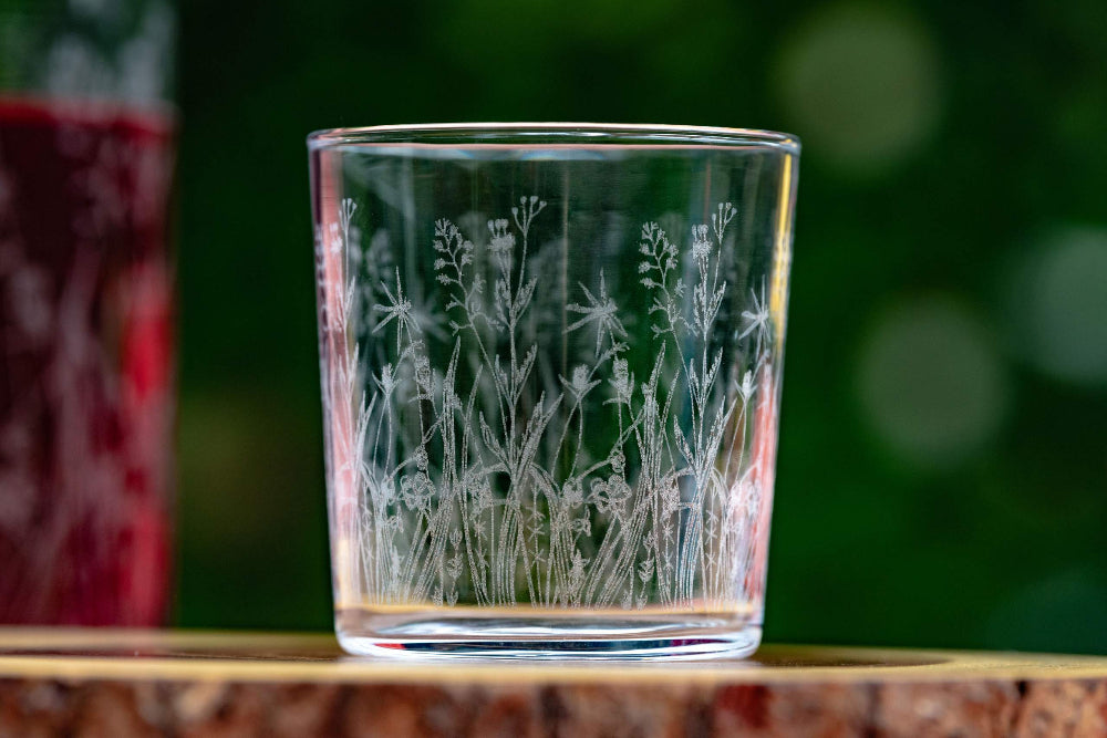 Glass tumbler with engraved floral design on a wooden surface with a blurred green background