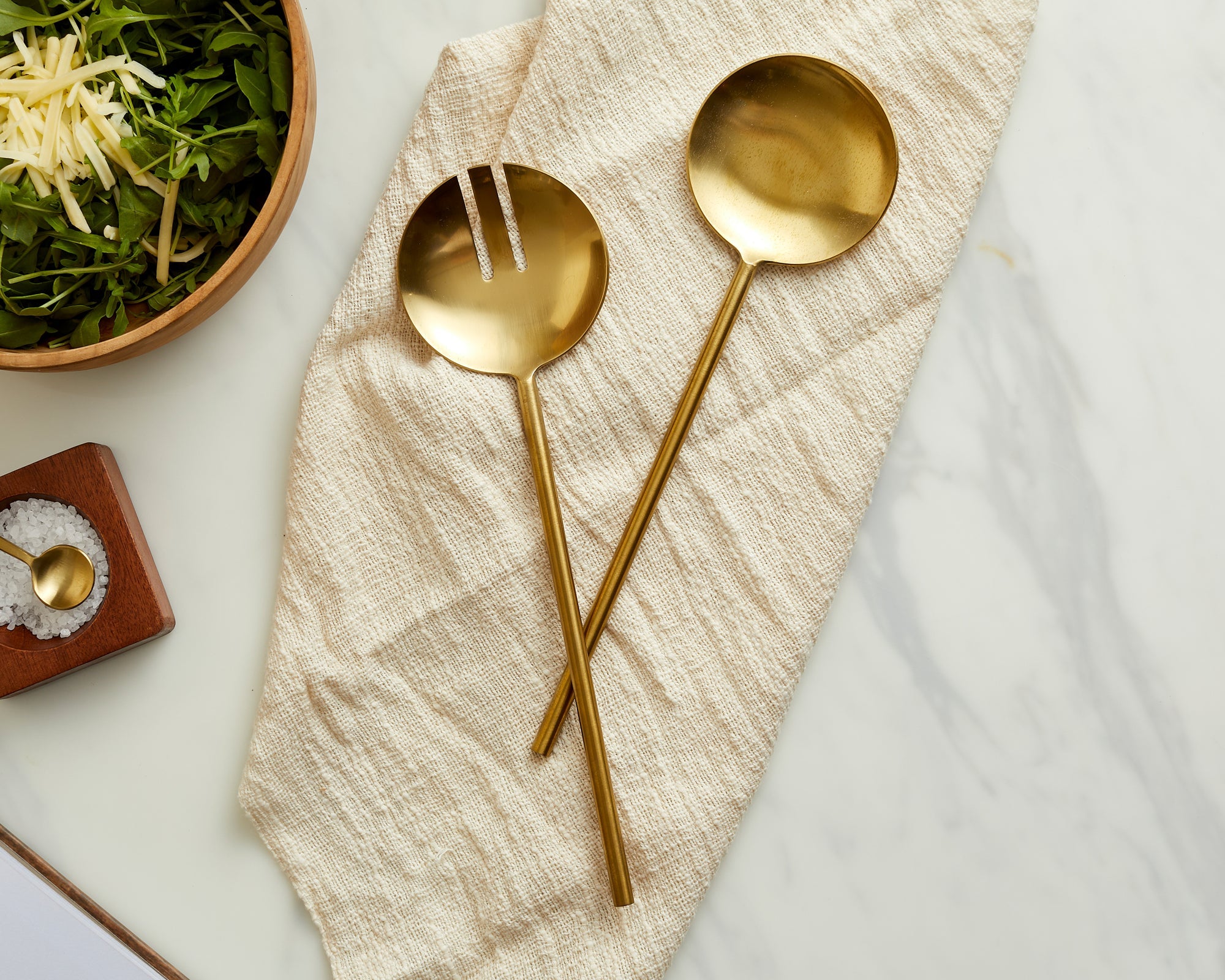 Two gold spoons on a textured beige napkin with a bowl of salad in the background.