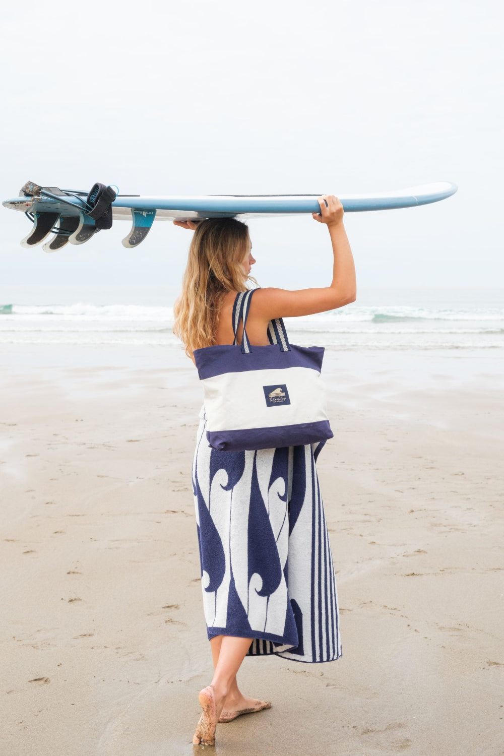 Woman carrying a surfboard and a tote bag with The Cornish Surfer logo on a beach.