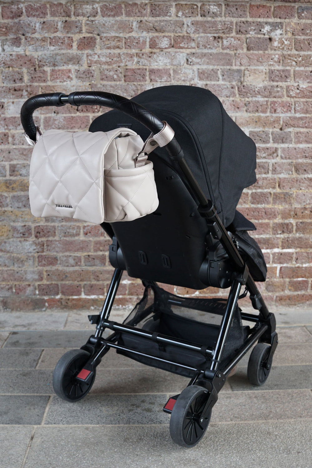 Black stroller with a Oyster pink coloured quilted bag against a brick wall.