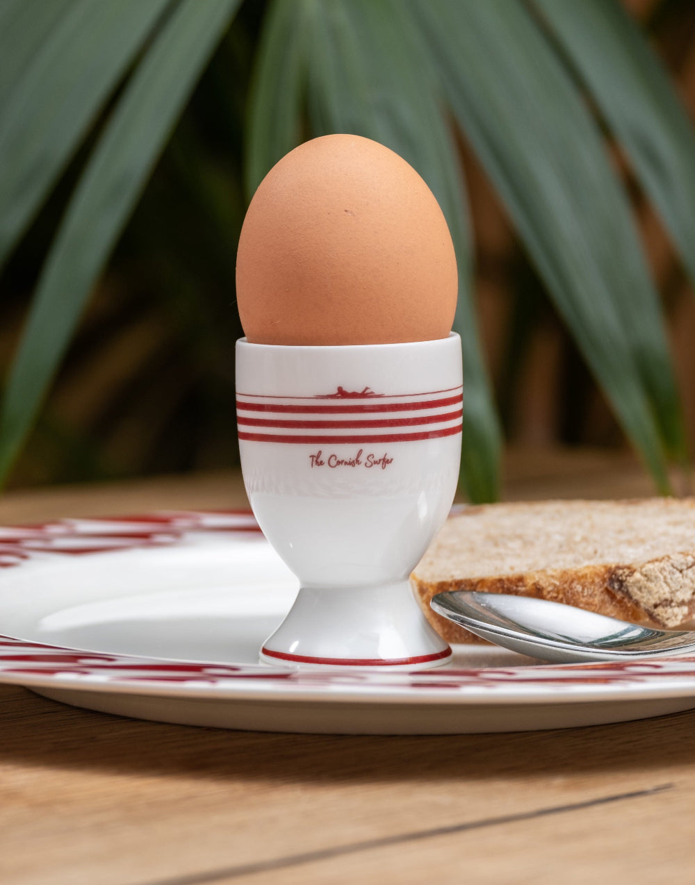 Brown egg in a white egg cup with red stripes on a plate with bread and a spoon.