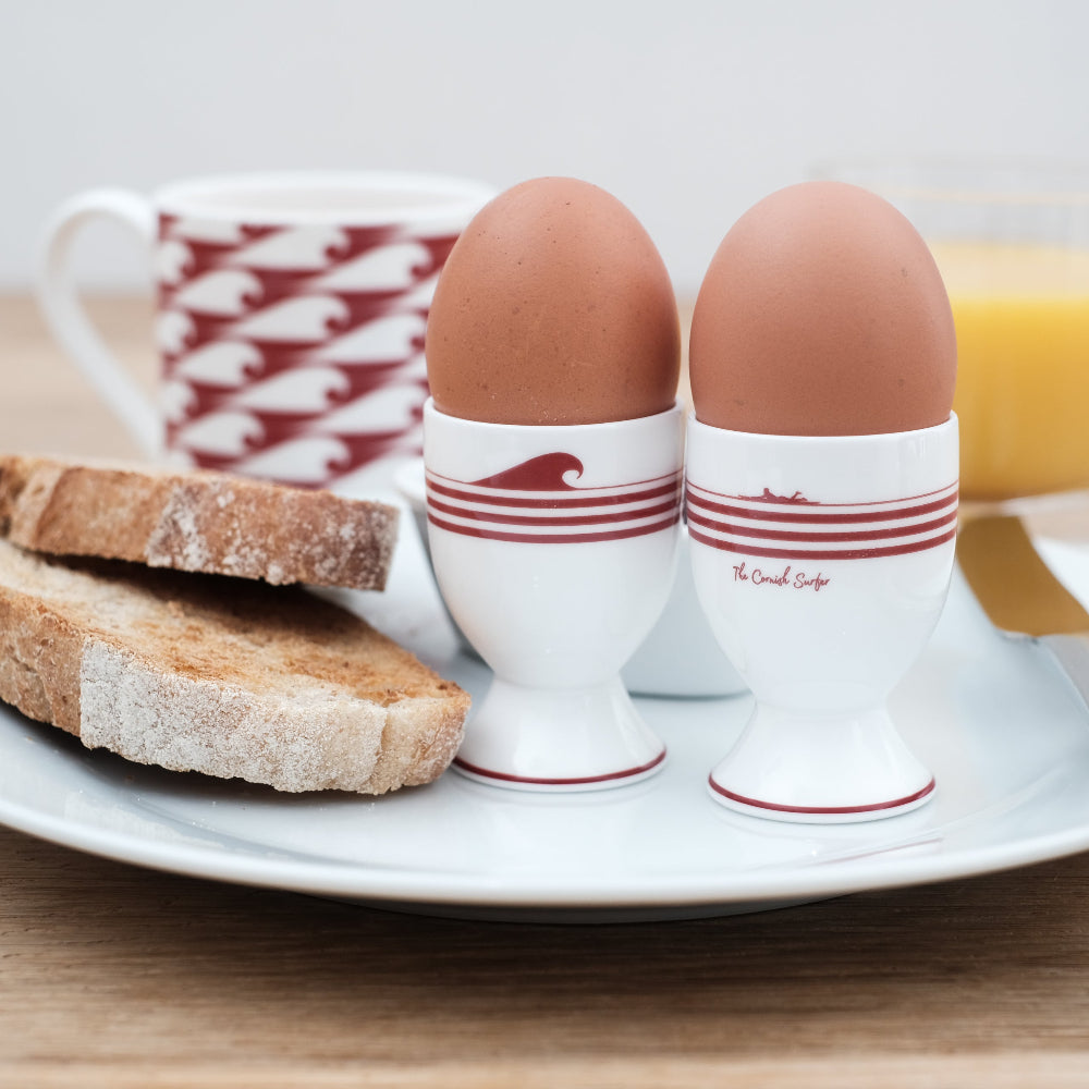 Two brown eggs in white egg cups with red stripes on a plate with toast and a cup of coffee.