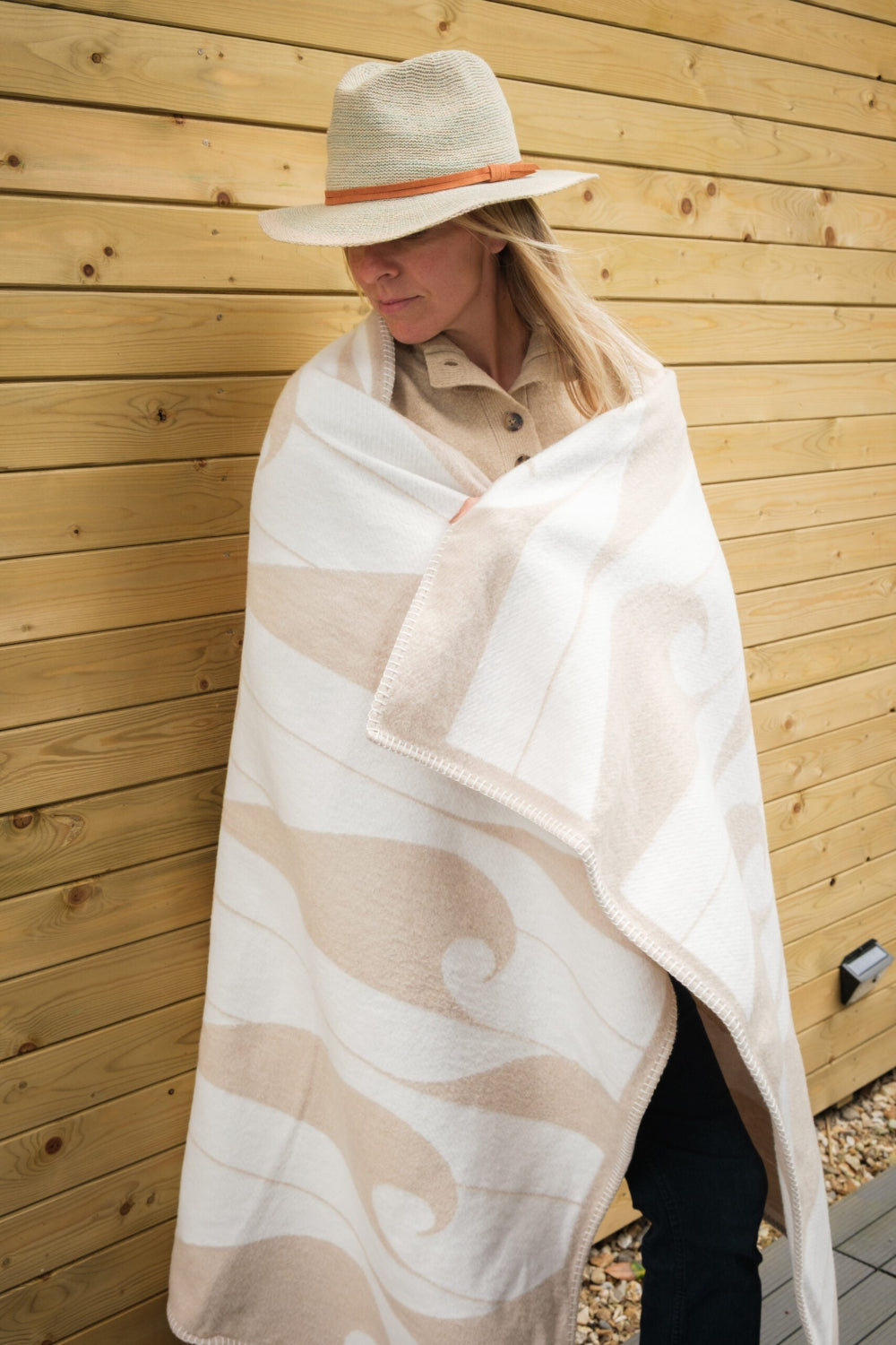 Person wrapped in a patterned blanket against a wooden wall with hat