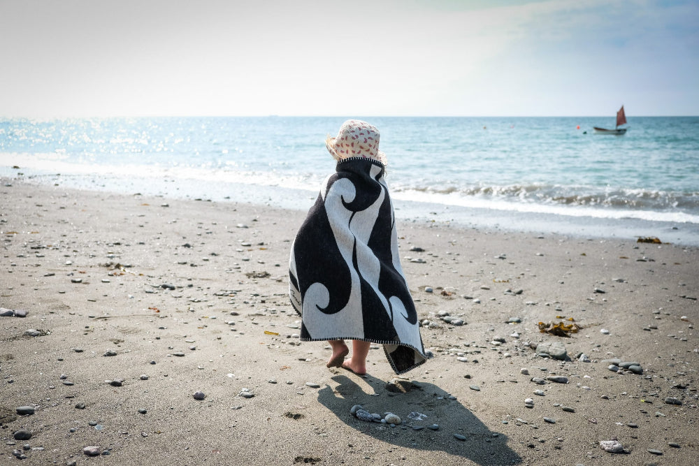 Person wrapped in a black and white patterned towel on a sandy beach with ocean in the background