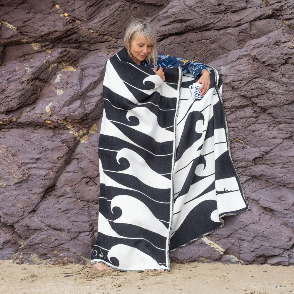 Person wrapped in a black and white patterned blanket on a rocky beach.