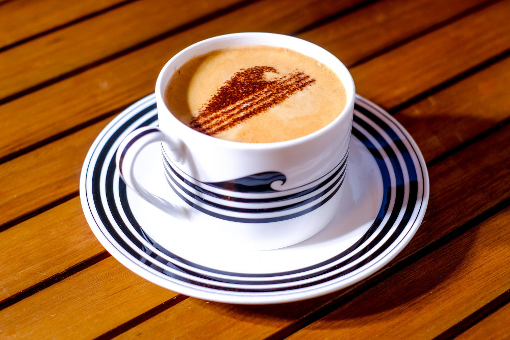 Cappuccino with wave chocolate sprinkles on top in a white cup with a blue stripe, placed on a matching saucer on a wooden table.