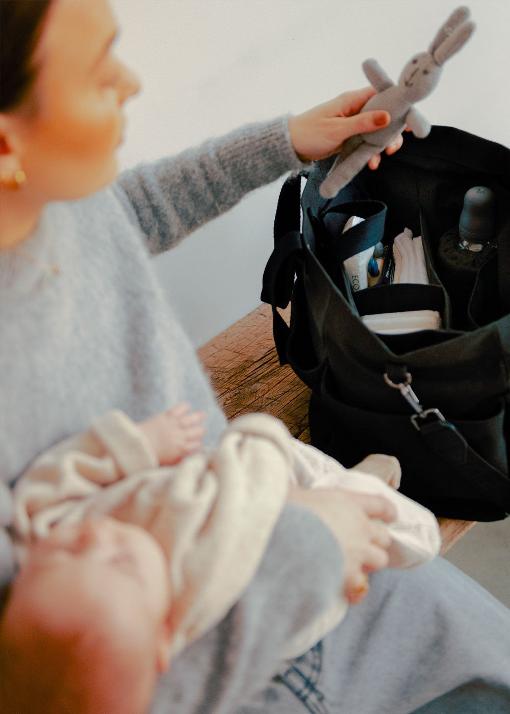 Person holding a baby and a stuffed animal next to the Tiba + Marl black Tote Canvas bag on a wooden bench.