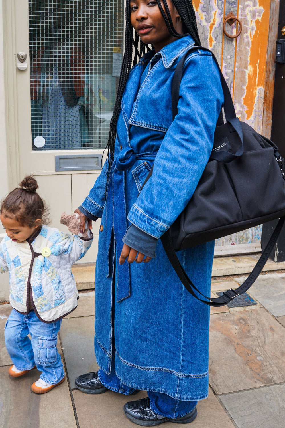 Woman in a blue denim dress holding a black bag, standing next to a child in a floral jacket on a street.