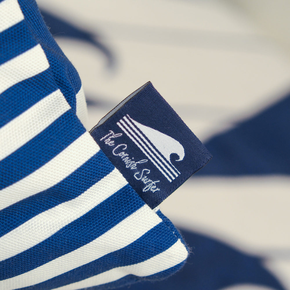 Close-up of a blue and white striped cushion with a brand label 'The Cornish Surfer' on a blurred background