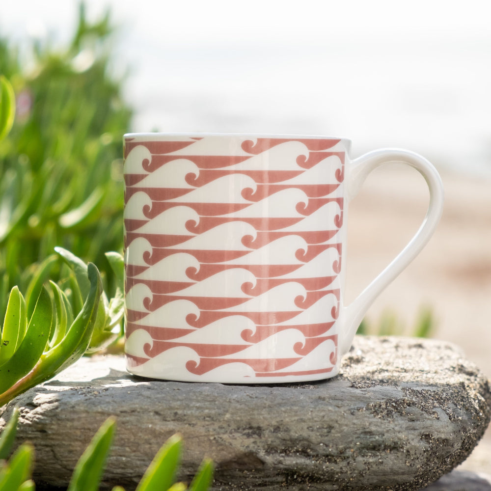 White mug with red and white wave pattern on a rock with greenery in the background