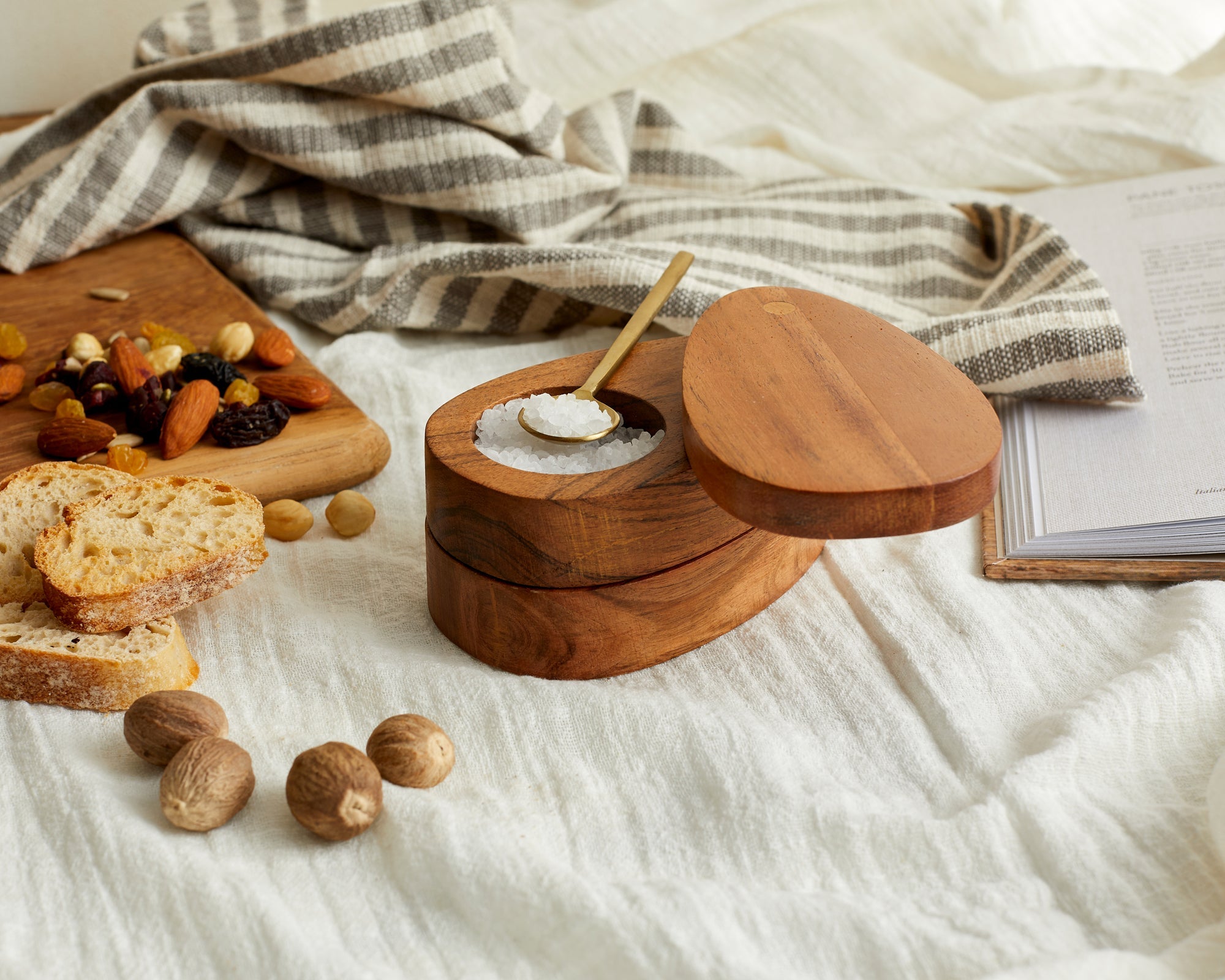 Wooden salt cellar with a spoon on a white surface with bread and nuts.