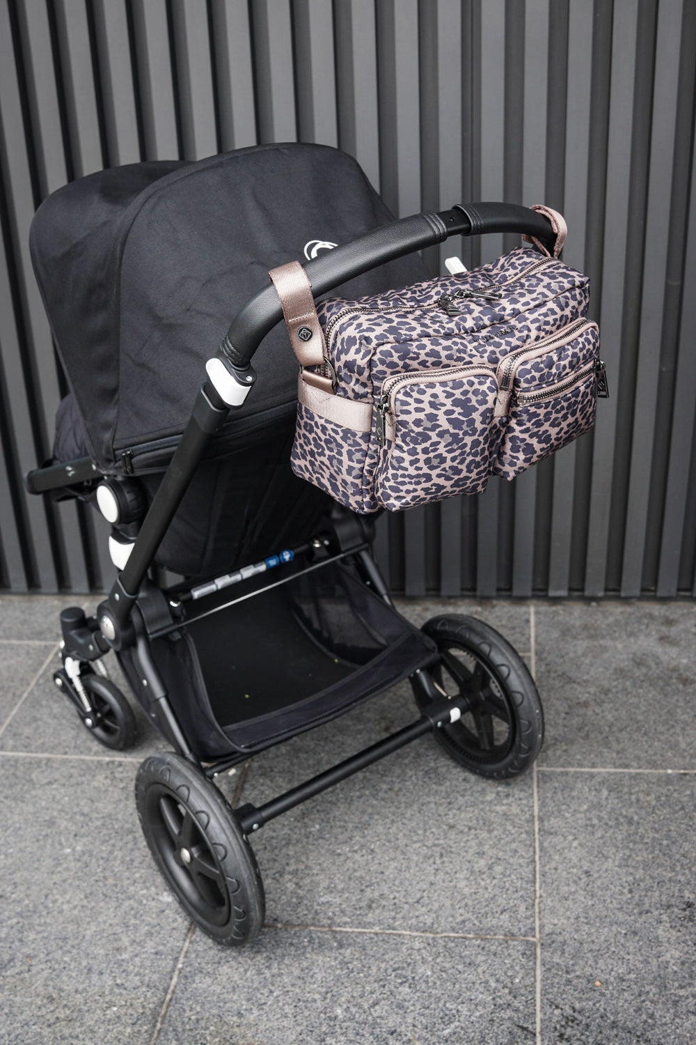 Black stroller with a leopard print bag attached to baby buggy handles on a tiled floor against a black wall.