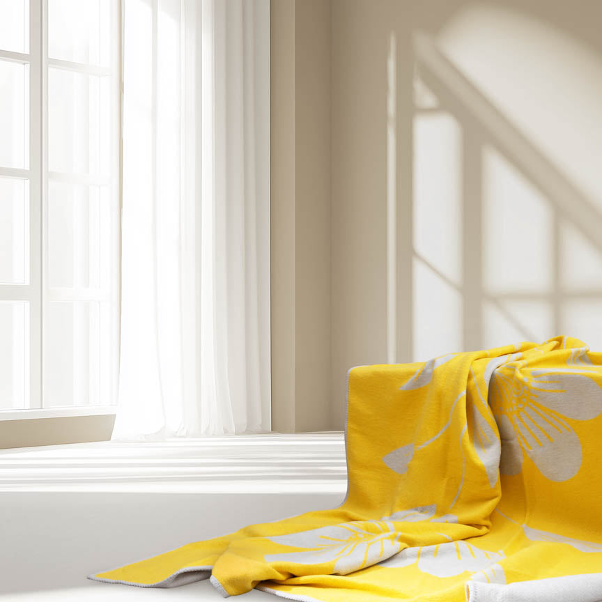 Yellow blanket with floral patterns draped over a surface near a window with white curtains.