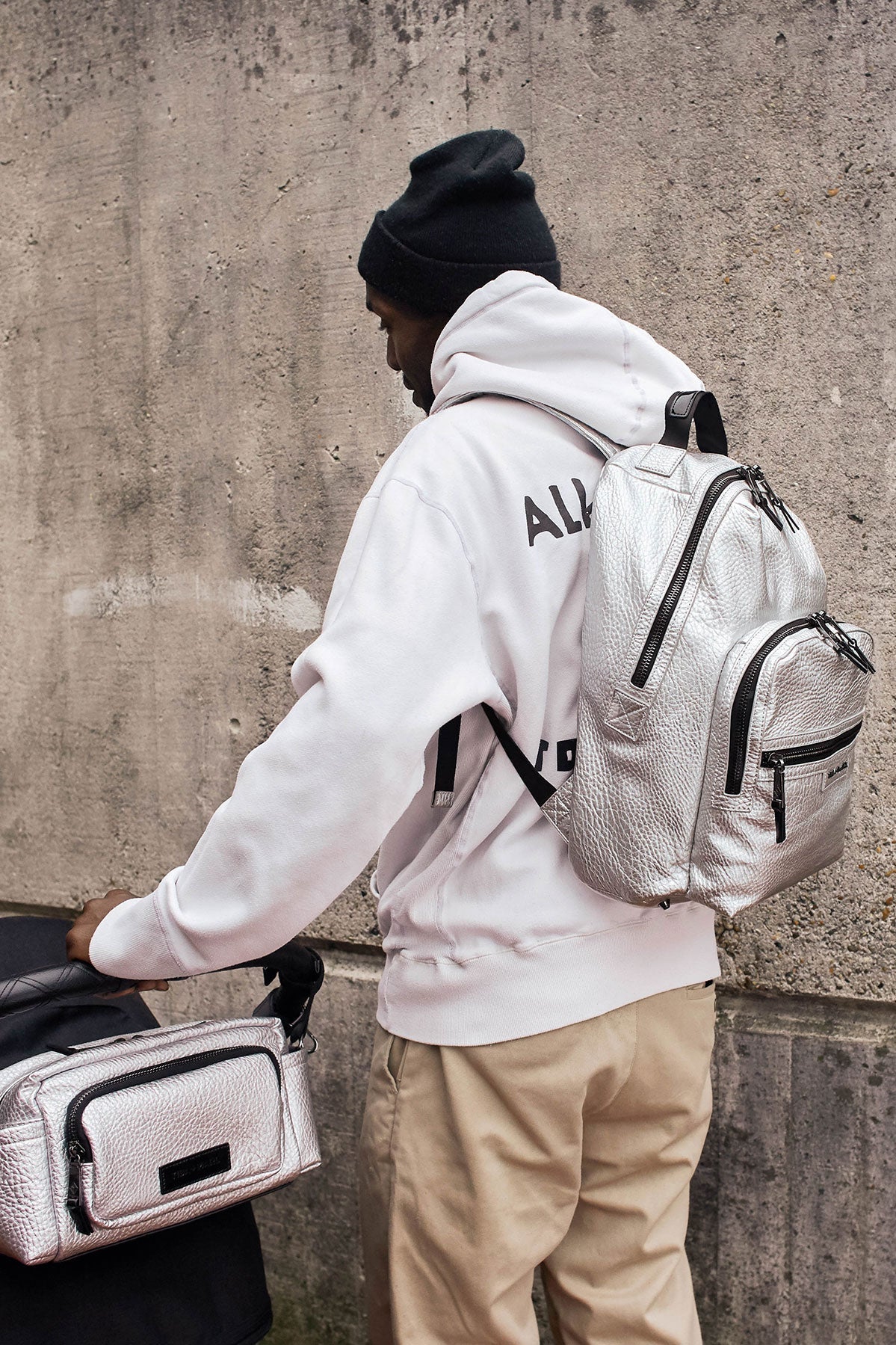 Person wearing a white hoodie, holding a matching backpack against a concrete wall.