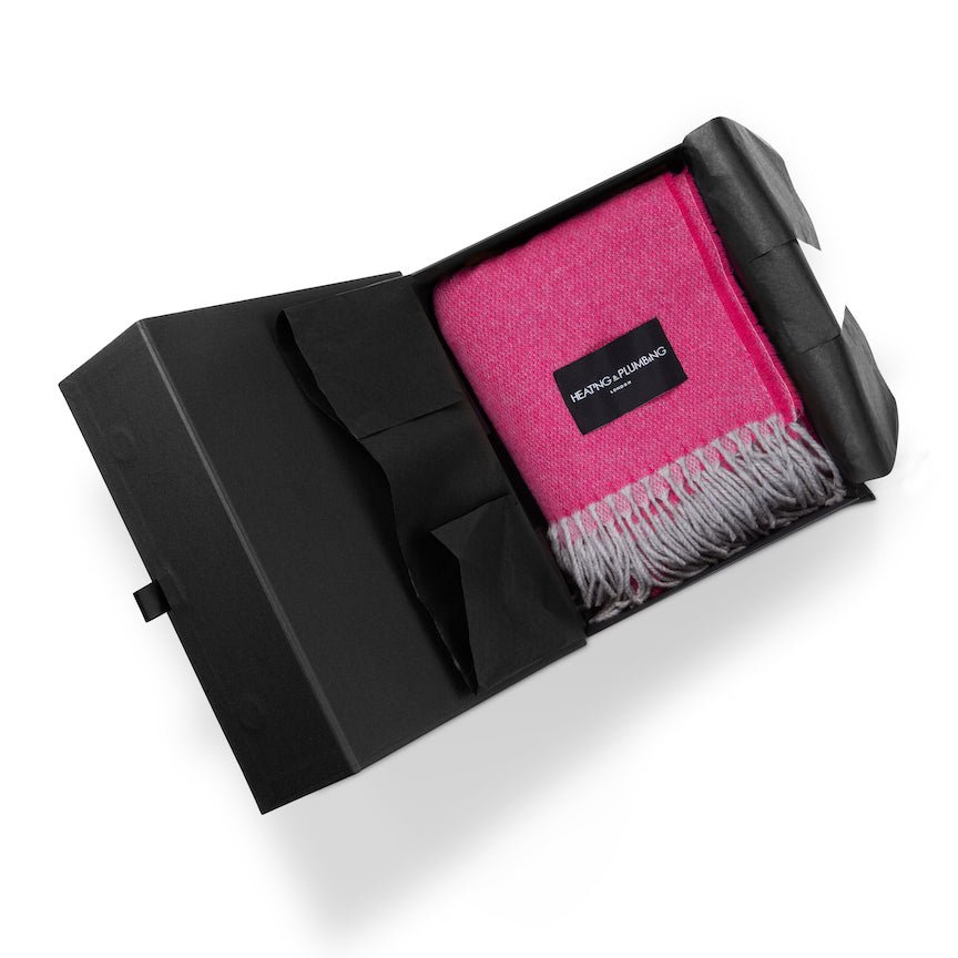 Pink blanket with grey fringe in a black box, featuring a brand logo.