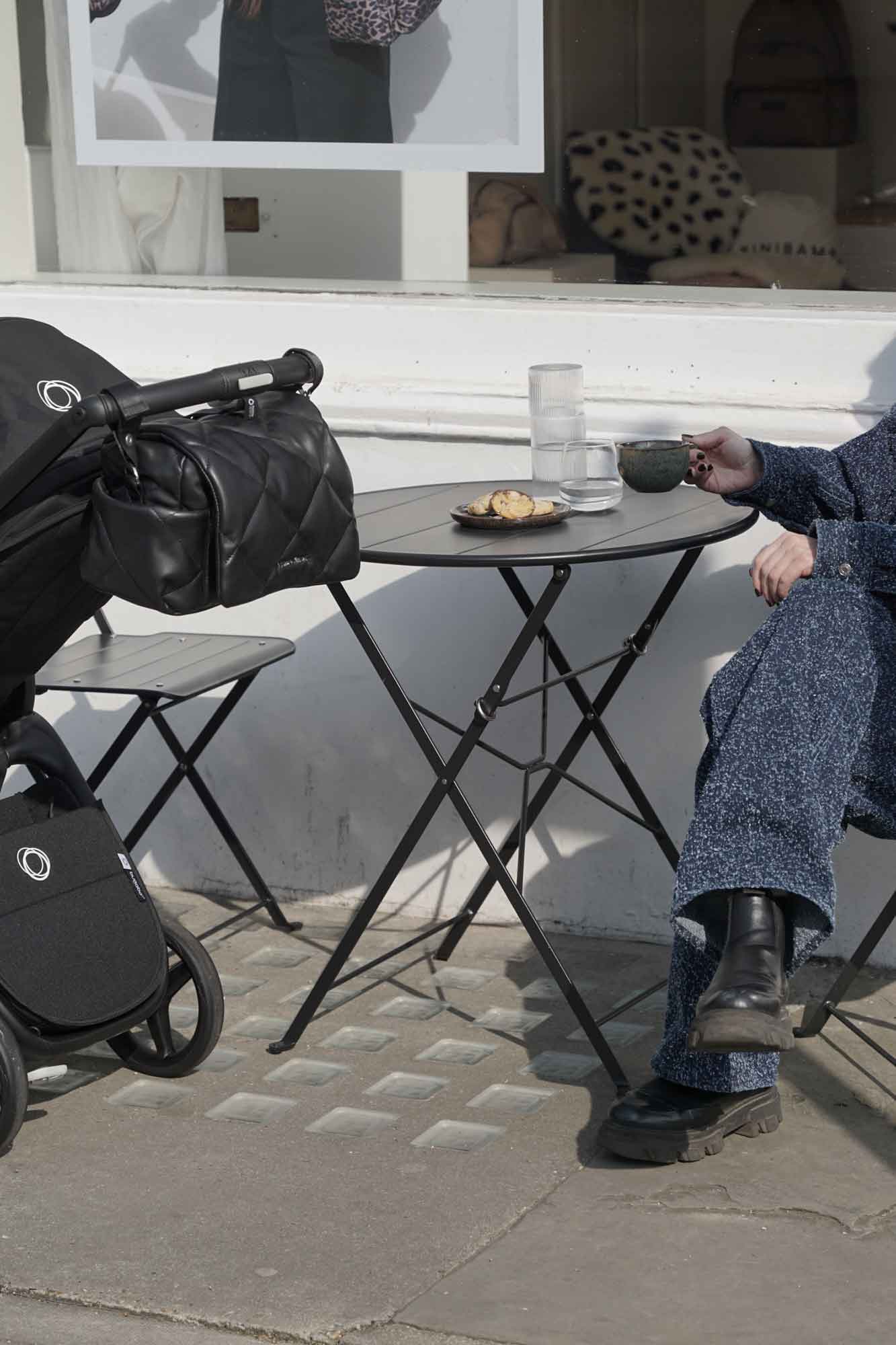 Person sitting at a table with a coffee cup with a stroller nearby on a stone pavement.