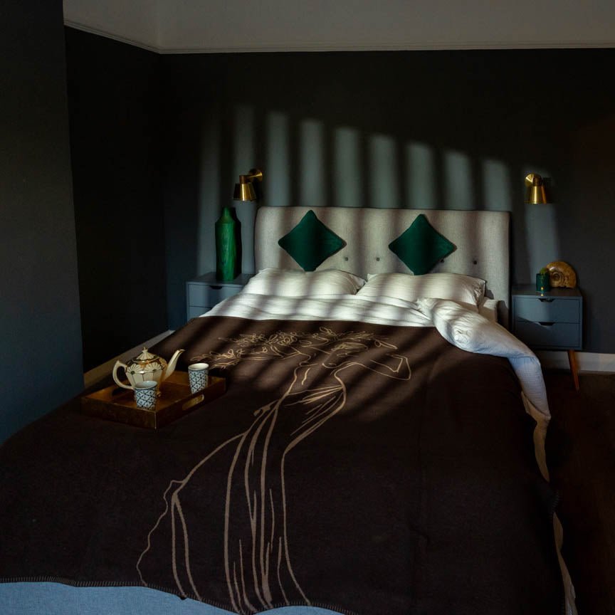 Bedroom with a bed featuring a dark brown blanket and green pillows, with a tray of tea on a nightstand.