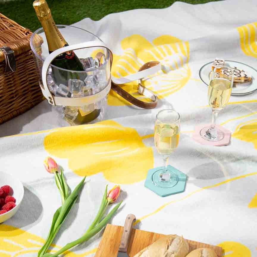 Picnic setup with yellow floral-themed blanket, wine, bread, and flowers on a grassy background.