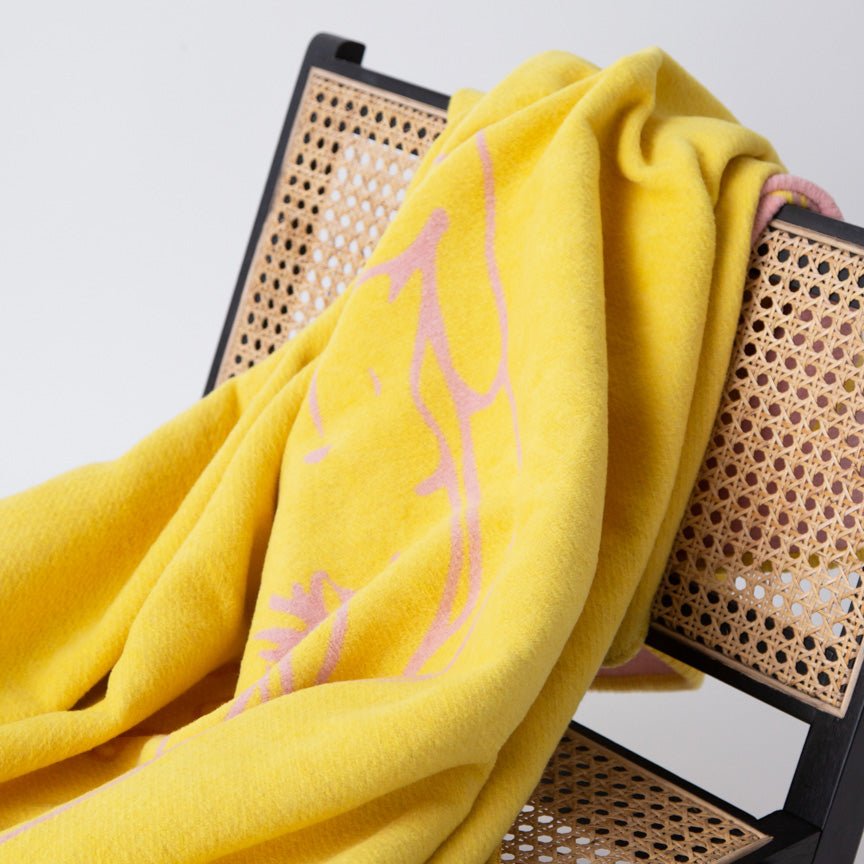 Yellow blanket with pink patterns draped over a wicker chair against a white background