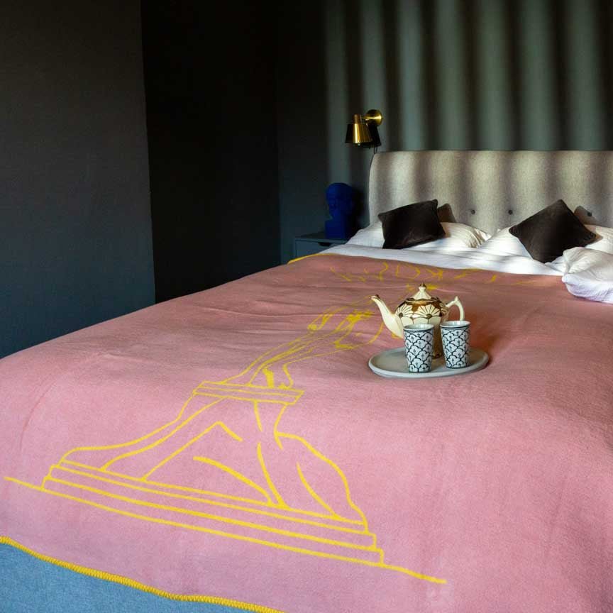 Bed with pink blanket featuring yellow line art, teapot, and cups on a dark background